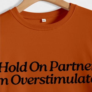 Orange sweatshirt featuring 'Hold On Partner I'M Overstimulated' text print with crew neck