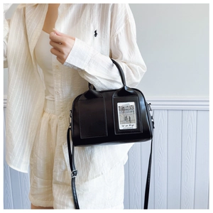 Detail of a black matte shell bag worn with a chic outfit against a wooden wall