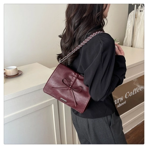 Close-up of a burgundy crossbody bag with a bow detail and chain strap worn by a model