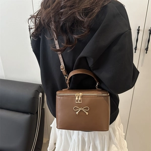Brown box-shaped crossbody bag with metal bow detail worn by model in home setting