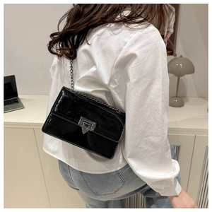 Detail of black patent leather crossbody bag with metal chain strap worn by a model