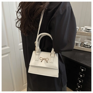 Close-up of white leather handbag featuring a decorative gold metal bow on the front flap