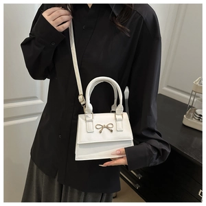Close-up of a white leather handbag featuring a delicate gold metal bow and rigid handle
