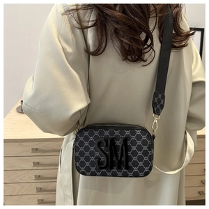 Detail of grey camera bag with SM logo, wide strap, and monogram pattern worn by a model