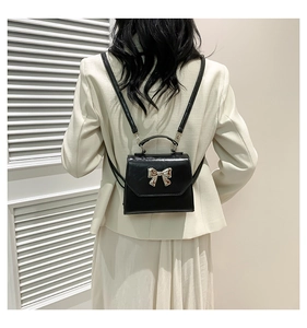 Back view of a black mini backpack with a gold bow buckle on a model in a minimalist room