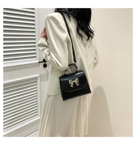 Model wearing a black leather crossbody bag with a silver bow clasp in a minimalist room