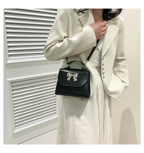 Détail d'un sac bandoulière en cuir noir avec nœud argenté porté sur une robe blanche