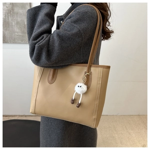 Beige leather tote bag with brown handles and plush charm shown on grey coat model