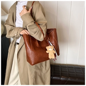 Brown leather tote bag featuring a plush bear pendant worn by a model in a beige trench coat