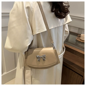 Detail of a beige crossbody bag with a silver bow accent worn on a white trench coat