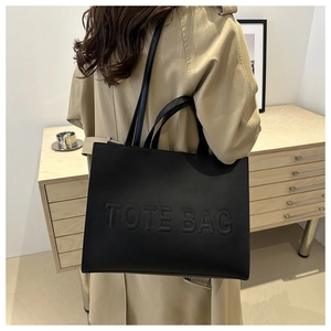 Detail of embossed TOTE BAG lettering on black leather tote worn with a beige trench coat