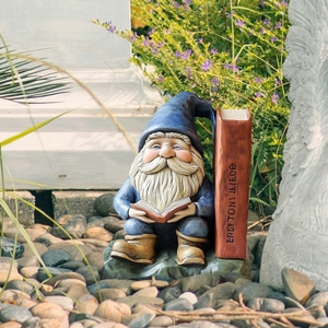 Detail of garden gnome reading a book figurine on stones among garden greenery