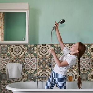 Close-up of a chrome shower head and hose held by a child in a vintage tiled bathroom