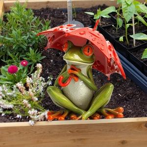 Detail view of a green frog statue holding a red umbrella in a garden flower bed