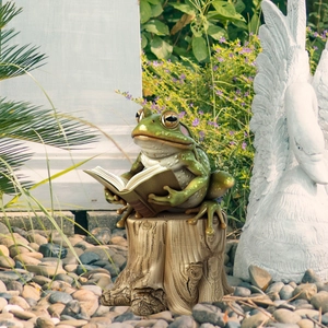 Detail of garden resin frog statue reading a book on a stump among river stones