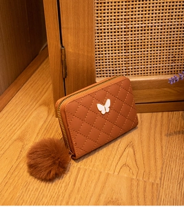 Detailed view of a brown quilted zip purse with butterfly metal logo and a plush pompom charm