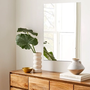 Modern frameless rectangular wall mirror above a wooden dresser in a bright living room