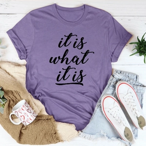Flachgelegtes lila Baumwoll-T-Shirt mit 'it is what it is' Spruch auf weißem Holzboden