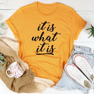 Flatlay eines orangen 'it is what it is' T-Shirts auf Holz mit lässigen Accessoires