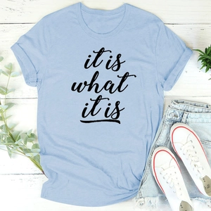 Flatlay des hellblauen 'it is what it is' T-Shirts mit Denim-Shorts auf weißem Holz