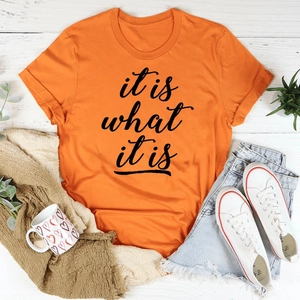 Flatlay eines orangen T-Shirts mit dem kursiven Aufdruck 'it is what it is' auf Holz