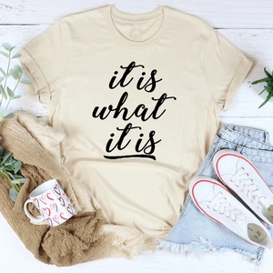 Beiges Baumwoll-T-Shirt mit it is what it is Schriftzug auf weißem Holz-Hintergrund