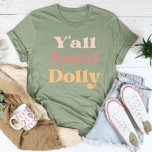 T-shirt en coton vert sauge avec motif vintage Y'all Need Dolly sur fond en bois blanc