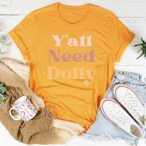 T-shirt orange à col rond avec texte rose Y'all Need Dolly posé sur fond de bois blanc