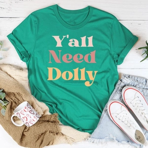 T-shirt vert en coton avec texte Y'all Need Dolly sur fond en bois blanc, style plat