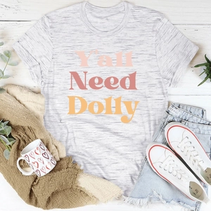 T-shirt gris à plat avec texte Y'all Need Dolly en dégradé sur fond de bois blanc