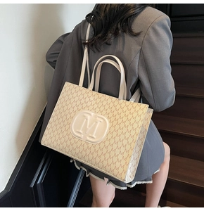 Detail of a beige tote bag with M letter pattern carried by a model on wooden stairs