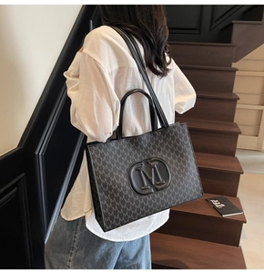 Détail d'un grand sac cabas noir avec motif M porté par un mannequin dans un décor moderne