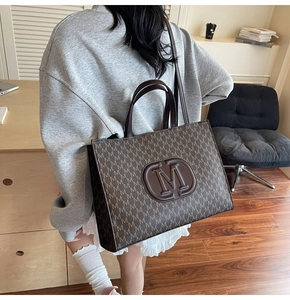 Brown monogram tote bag with M logo shown on model's shoulder in office setting