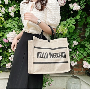 Linen tote bag detail with HELLO WEEKEND print and black handle against floral brick wall