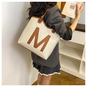 Detail of cream woven tote bag featuring brown leather trim and a large M patch