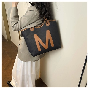 Detail of a black woven tote bag featuring a large brown M letter patch and faux leather handles