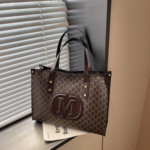 Large coffee tote bag with M-letter print and wide leather straps in natural sunlight