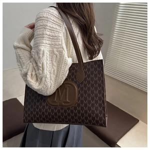Detail of dark brown tote bag with M-pattern print and leather logo on shoulder