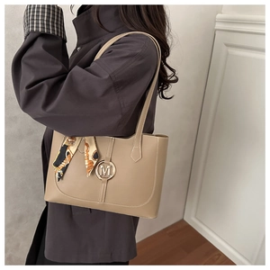 Detail of khaki leather tote bag featuring a round metal logo and a silk scarf on the strap