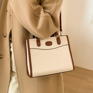Minimalist beige tote bag with brown trim and gold buckle worn by model against a warm wall