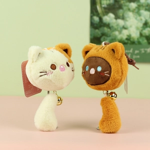 Close-up of two plush cat charms with golden bells and long fluffy tails on soft pastel background