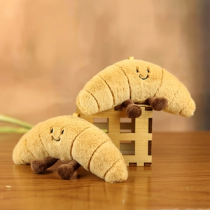 Cute plush croissant keychain with embroidered smiley face and tiny brown legs on wood