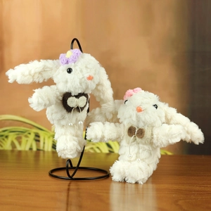 Cute white plush bunny keychain with knitted flowers and pearl bow on a warm wooden stand