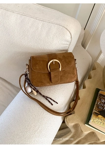 Brown suede shoulder bag with gold buckle and baseball charm on white sofa