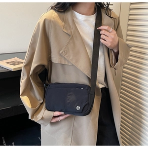 Detail of a black nylon crossbody bag with metal logo, worn with a beige trench coat