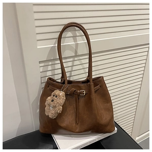 Brown leather tote bag with buckle detail and plush bear charm on a park bench