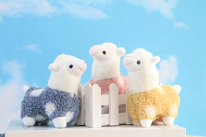Three plush alpaca toys in blue, pink, and yellow displayed on a white fence prop