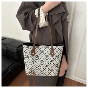 Detail of a beige tote bag with blue patterns, brown leather handles and gold M charm