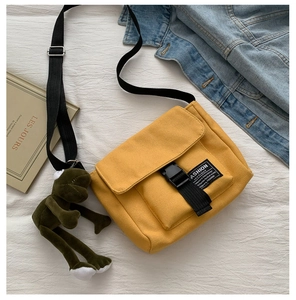Detail of yellow canvas messenger bag with black buckle and green frog plush charm