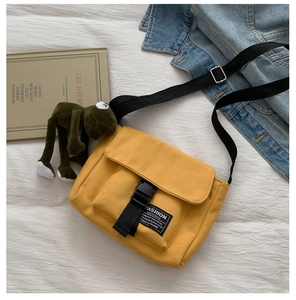 Flat lay of yellow canvas messenger bag with black buckle and plush frog pendant charm